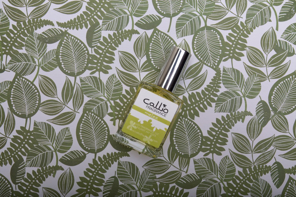Bottle of one-ounce Melee perfume lying on a white background with green leaves.